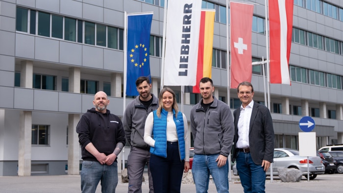 SICK and Liebherr employees standing in front of the Liebherr site building with European and national flags in the background